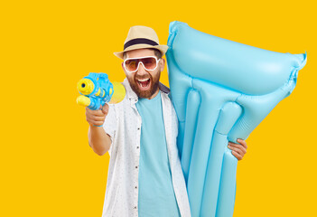 Happy cheerful man having fun on holiday. Bearded young guy in summer shirt, sun hat and glasses enjoying vacation, holding blue inflatable mattress and shooting from water gun on yellow background