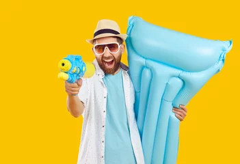 Foto op Canvas Happy cheerful man having fun on holiday. Bearded young guy in summer shirt, sun hat and glasses enjoying vacation, holding blue inflatable mattress and shooting from water gun on yellow background © Studio Romantic
