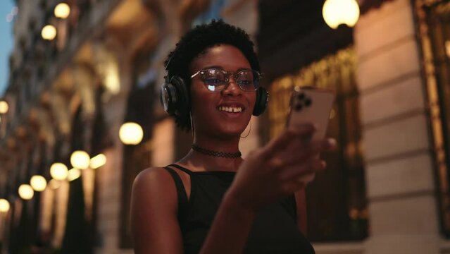 Happy smiling black female walk on evening city street, holding smartphone device that light face. Modern woman technology user enjoying favorite audio book in headphones while using app. Slow motion