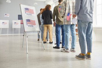 Large group of diverse people registering at polling station holding American flags in hands on election day. American voters standing in queue at vote center going to put ballots in voting booth. - obrazy, fototapety, plakaty