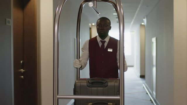 Young African American porter in uniform going through hotel corridor and pushing luggage cart