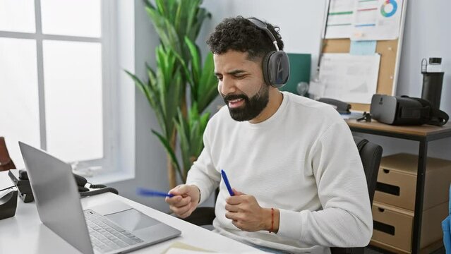 A bearded young man wearing headphones plays with pens as drumsticks in a modern office setting.