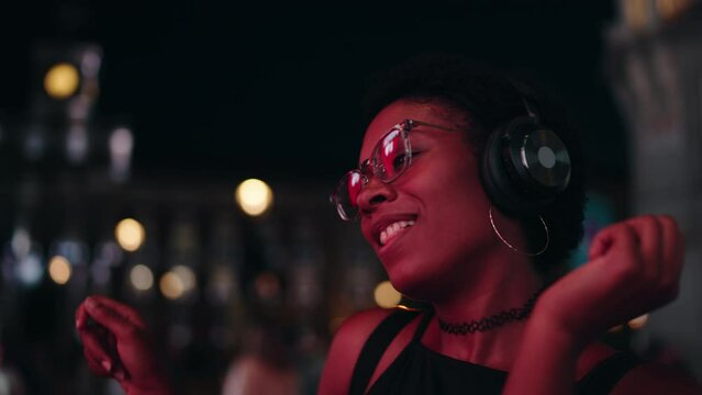 Expressive carefree young woman celebrates night through dance and song, her passion illuminated by urban glow. Relaxed happy black female enjoying listening to music in headphones. Slow motion
