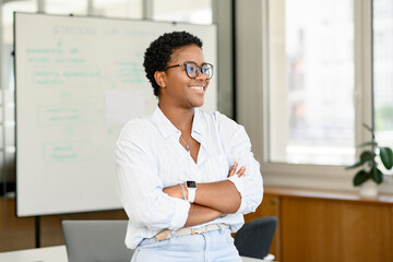 African-American businesswoman standing in modern office near desk with a laptop and looking away....