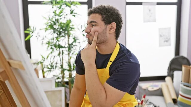 Thoughtful man with beard in apron painting on canvas in bright art studio