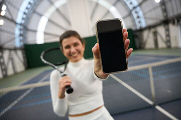 Smiling fit woman player showing blank phone screen on tennis court