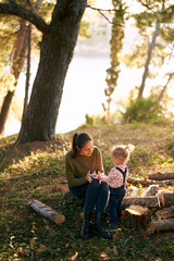 Little girl stands near her mother sitting on a stump in the forest and plays with a toy