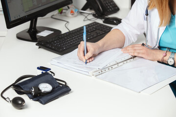 Hands of general practitioner filling paper medical records. Doctor in white coat doing paperwork at workplace with laptop, writing notes, preparing documents, reports, prescription. Close up.