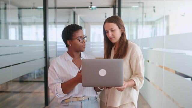 Two coworkers chatting during coffee break standing in glass hall of office building. African-american female employee explain something to colleague, sharing ideas