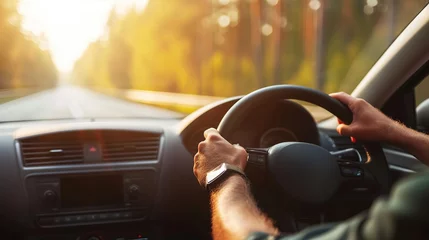 Fotobehang The image shows a person's hands on a steering wheel, driving a car on a sunny road surrounded by trees. © Oleksii