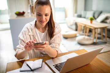 Young female freelancer working on laptop using smartphone at home