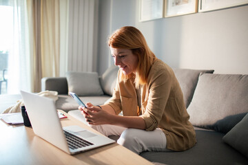 Smiling young woman on the couch using smartphone next to laptop