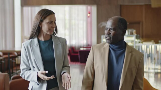 Medium shot of young Caucasian businesswoman speaking with mature African American colleague while walking together through hotel restaurant