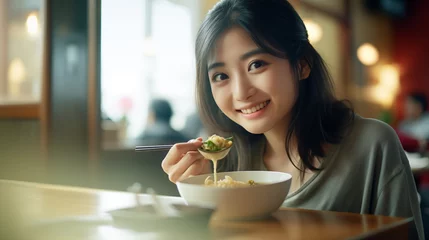 Gordijnen The image depicts a young Asian woman smiling while eating with chopsticks in a restaurant. © S photographer