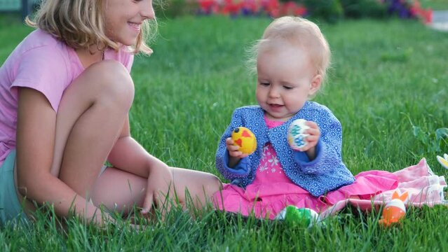 Adorable baby girl playing with Easter eggs while older sister sitting on a grass nearby