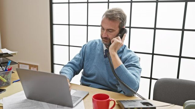 Middle-aged man with beard on phone, using laptop in office, showcasing work environment