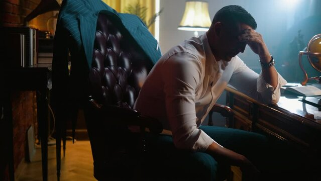 Exhausted businessman rests his head on his hand, sitting disappointed in an armchair in a luxury office late at night, thinking about solving problems and solutions. He feels stressed, has a headache