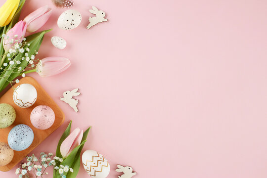 Celebrate Easter with style. Top view flat lay of colorful eggs, fresh tulips, easter bunnies, feathers on pastel pink background with advert zone