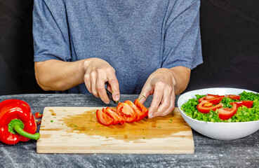 Female hands cutting tomato on cutting board. Blue t-shirt, black background. Salad preparation.