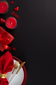 Sumptuous Valentine's Day repast. Top view vertical shot of plates, cutlery, hearts, present, red napkin, candles on black background with advert space