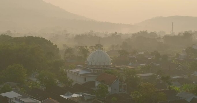 Aerial Forward Shot Of Mosque Amidst Residential Houses In Town By Mountains During Sunset - Java, Indonesia