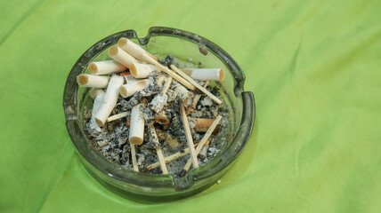 Fototapeta na wymiar Ashtray full of cigarette butts on green background. Unhealthy life and bad addiction. 