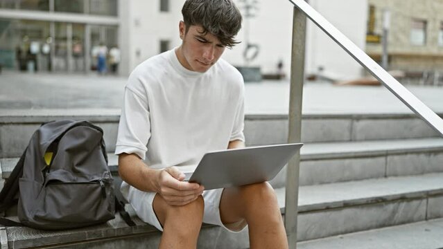 Cool young hispanic teenager, a smart university student engrossed in online study on laptop while sitting casual on stairs at campus, portraying relaxed concentration under city sunlight.