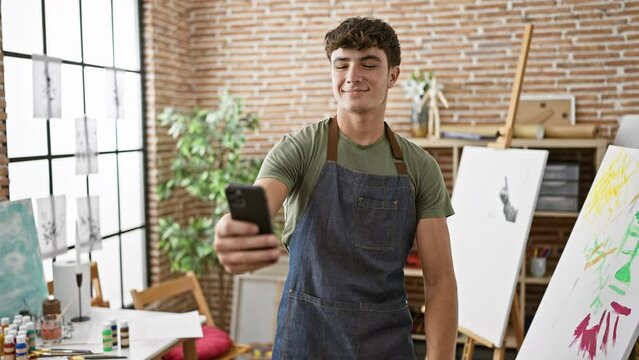 Confident young hispanic teenager artist, smiling, making a memorable selfie in his art studio, capturing his painting journey on smartphone