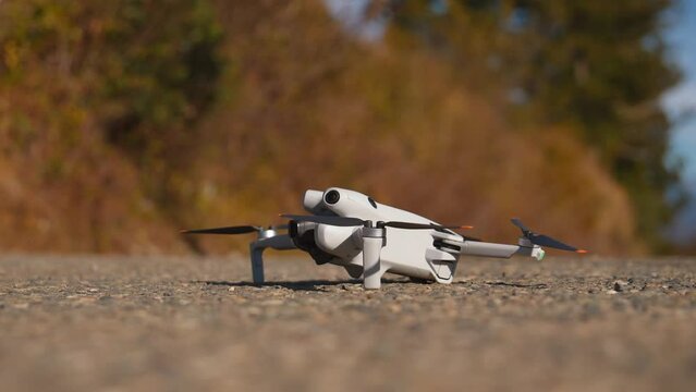 Small white mini drone taking off from surface of ground in autumn in park. It takes off from asphalt road against background of yellow autumn foliage.