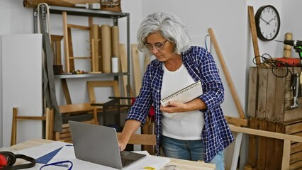 Mature woman with grey hair and glasses working on a laptop in a carpentry studio, surrounded by woodworking tools.