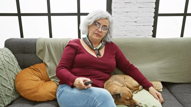 Elderly woman with grey hair and glasses lounging on a grey sofa with a tan dog indoors, remote in hand, expressing boredom