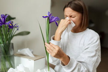 Allergic young woman holds iris flower, covers nose with paper tissue has runny nose, sneezes from...