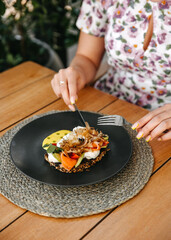 Person having breakfast, a poached egg on toast with salmon and avocado, garnished with herbs.