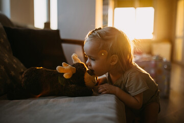 A toddler kissing a plush reindeer on nose, on a sofa, in a warmly lit room.