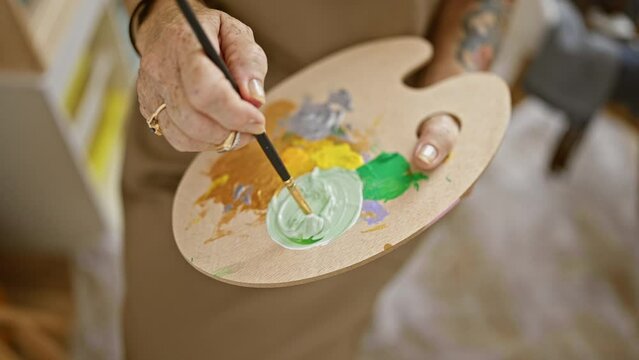 Senior woman artist's hands joyfully mix colors at art studio while mastering her painting hobby