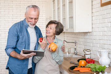 Senior happy couple grandparents spouses cooking together, helping with finding recipes for culinary dish, preparing food meal dinner lunch vegetable salad at home kitchen