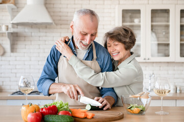 Cheerful old senior couple grandparents spouses hugging embracing cuddling while helping prepare cook romantic dinner lunch at home kitchen