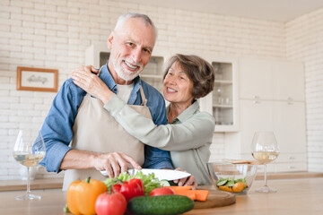 Ideal happy senior couple. Old elderly wife hugging embracing cuddling with husband, helping him prepare dinner, cooking vegetable salad together