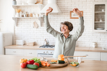 Active european senior old elderly woman grandmother dancing and singing while cooking preparing meal food dinner lunch vegetable salad at home kitchen