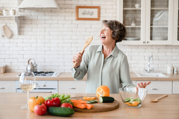 Happy active old senior elderly woman grandmother singing and using spoon as a microphone while cutting vegetables for salad, cooking at home kitchen - obrazy, fototapety, plakaty