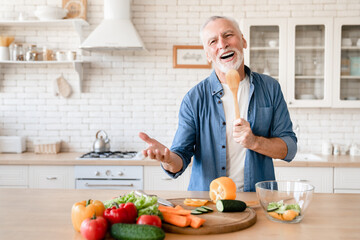 Funny cheerful caucasian senior old elderly man grandfather singing and dancing while cooking preparing meal food dinner vegetable salad at home kitchen