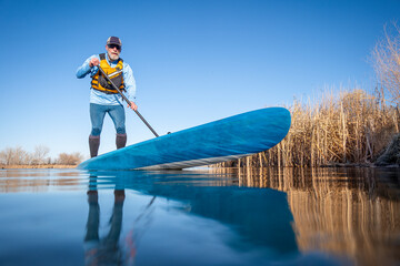 senior male paddler is paddling  a stand up paddleboard on a calm lake in spring, frog perspective...