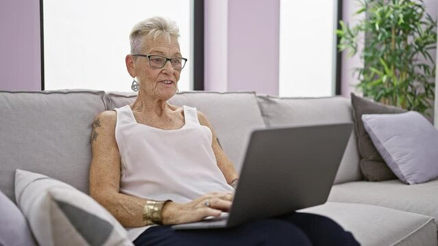 Confident grey-haired senior woman, smiling and enjoying restful moment on sofa at home, engaging with technology using laptop for relaxing video call