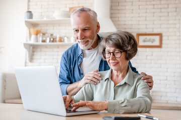 Active seniors concept. Caucasian old elderly senior wife and husband, couple spouses grandparents using laptop for e-banking, online shopping, remote work and telehealth at home kitchen - obrazy, fototapety, plakaty