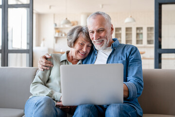 Fototapeta premium Caucasian senior happy old elderly grandparents couple spouses hugging embracing cuddling together while watching movie film series on laptop at home