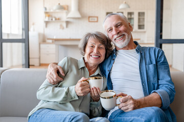 Cheerful old senior happy couple grandparents spouses drinking hot beverage tea coffee having breakfast while looking at camera sitting on the sofa, embracing and cuddling at home