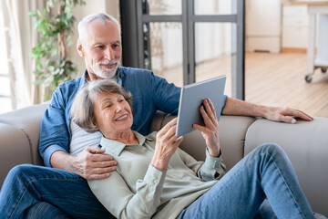 Relaxed cheerful old senior couple spouses grandparents watching movie film series, scrolling social media online, using digital tablet for online shopping at home together