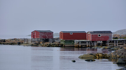 Three red wood boathouses sit on water at  the edge of the rocky shoreline on Fogo island in Newfoundland-Labrador
