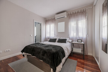 A bedroom with a double bed with a built-in wardrobe with white wooden sliding doors covering one wall and an air conditioner above the bed and an access door to a toilet