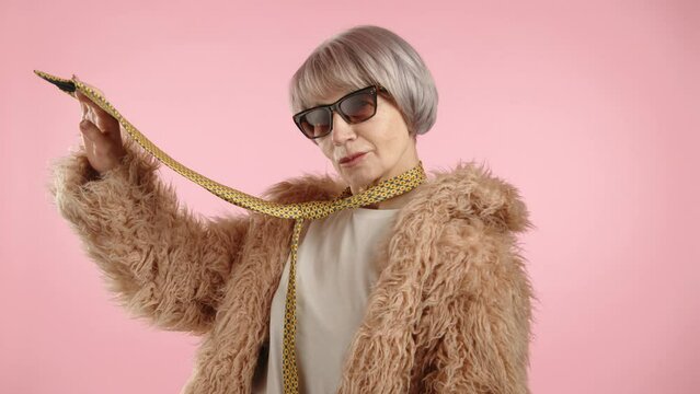 A woman in a fur coat with a necktie elegantly poses in front of the camera, playfully holding the tie in her hand. She has short blonde hair and wears sunglasses. The background is pink. 8K RAW.